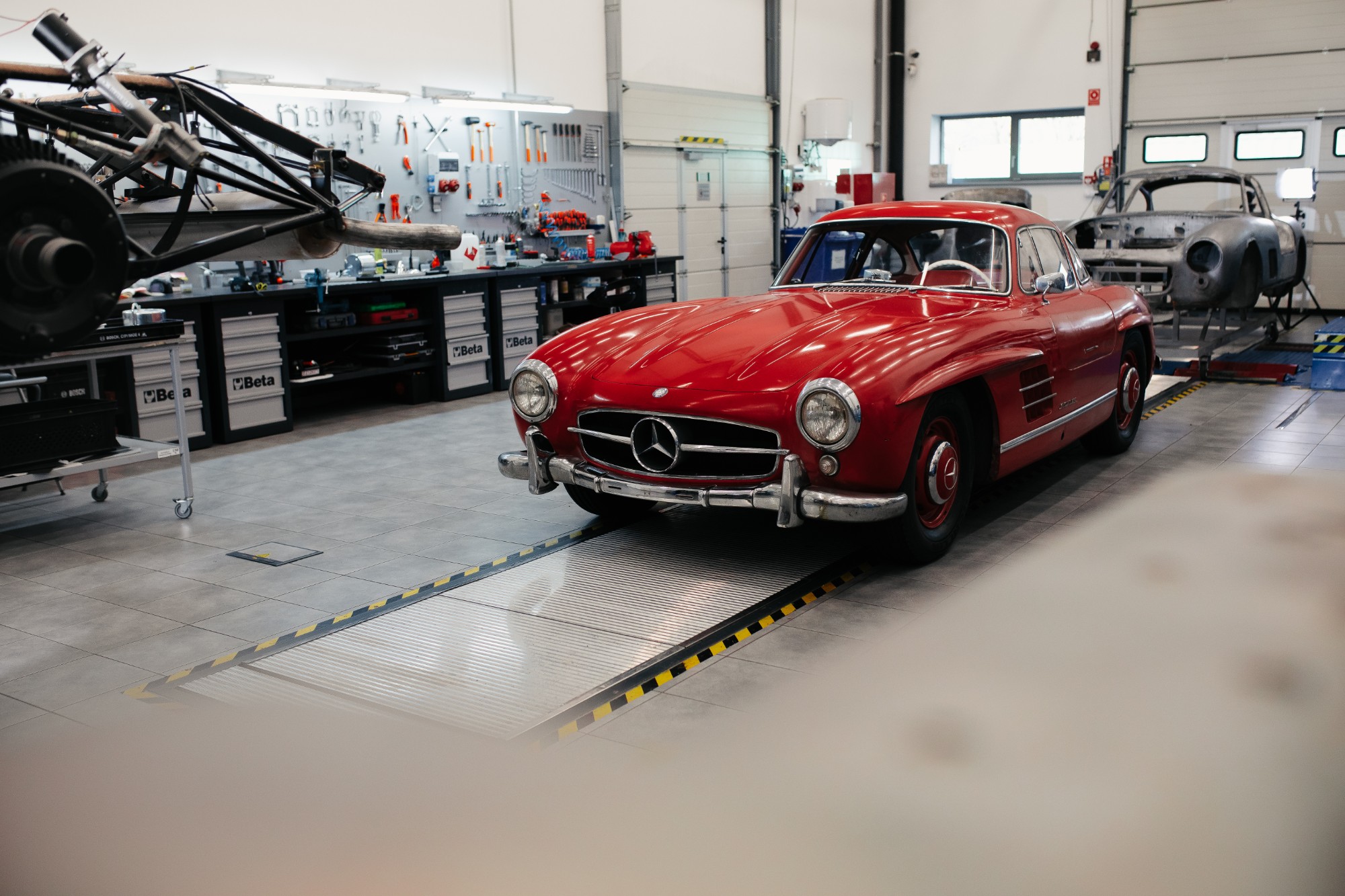 Red 300 SL Gullwing coupé in the Motor Classic workshop, with a bare-metal body on a frame in the background