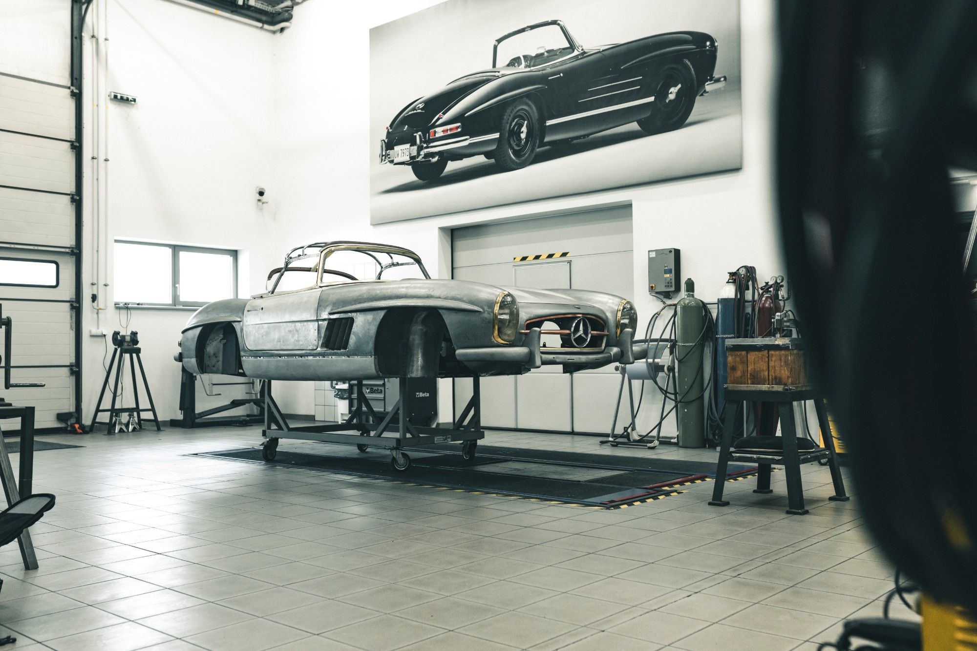 Bare-metal 300 SL Roadster body on a stand in the workshop, under a large photo of a black roadster