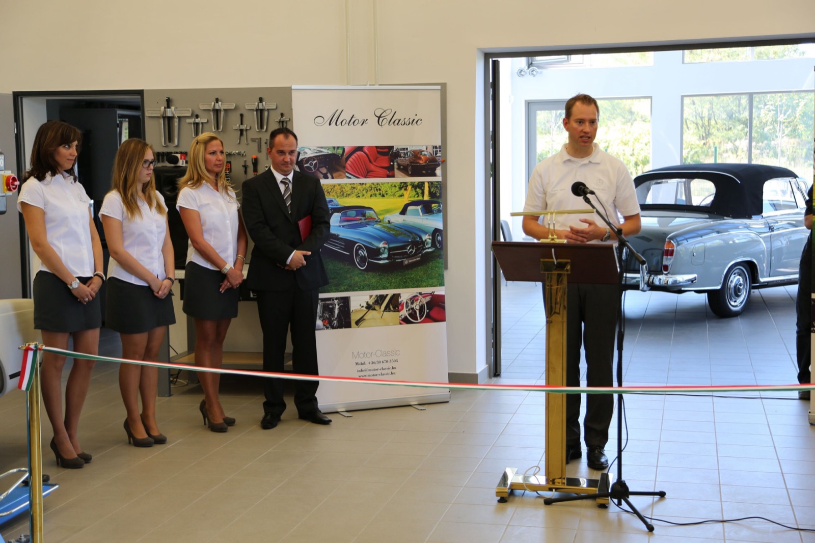 Speeches at the opening of the new workshop hall, with the ribbon in the national colours and a classic Mercedes-Benz cabriolet in the background