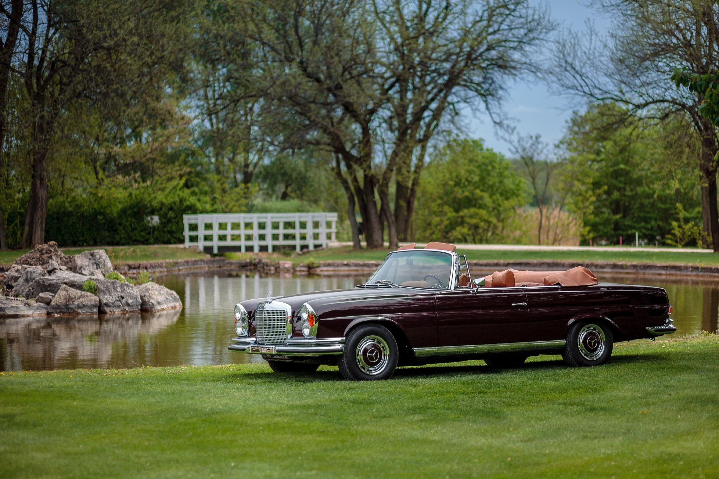 Maroon Mercedes-Benz W111 convertible on a lawn by a lake