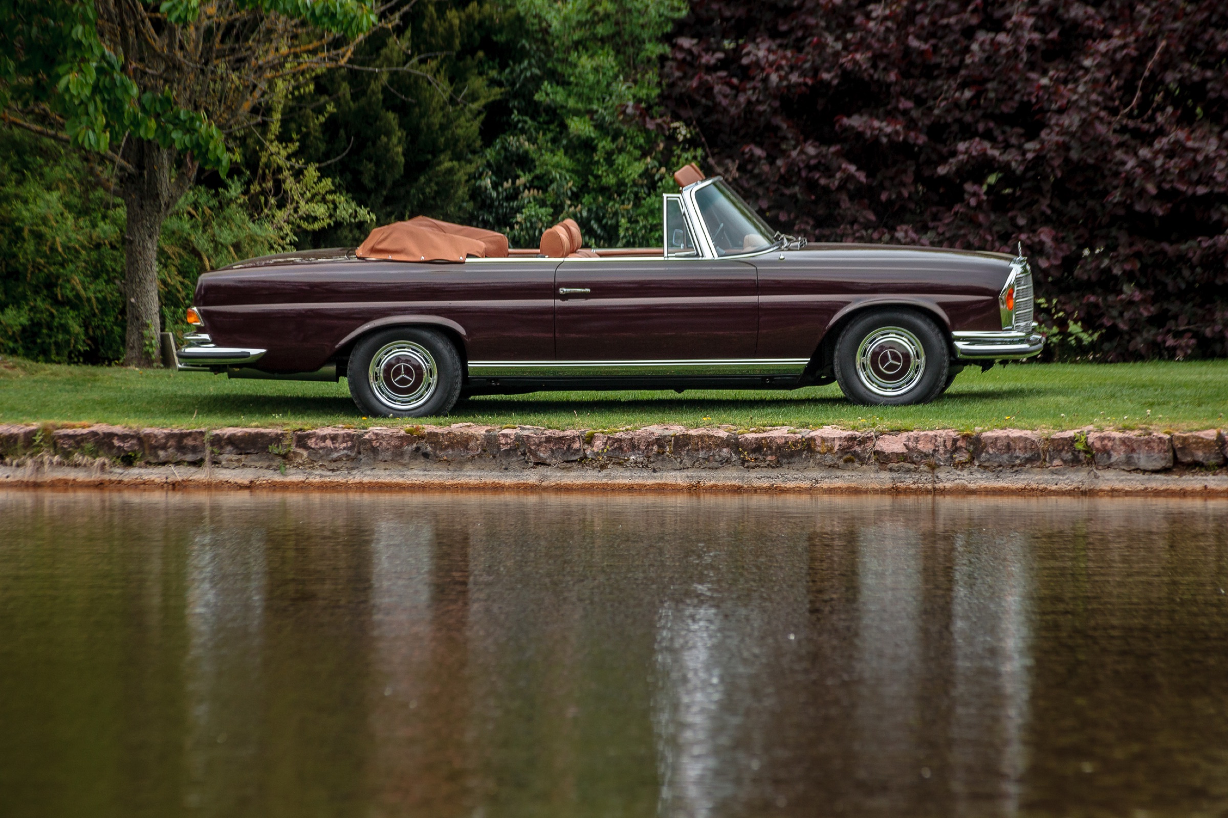 Side view of a maroon W111 convertible with the soft top folded, beside a pond